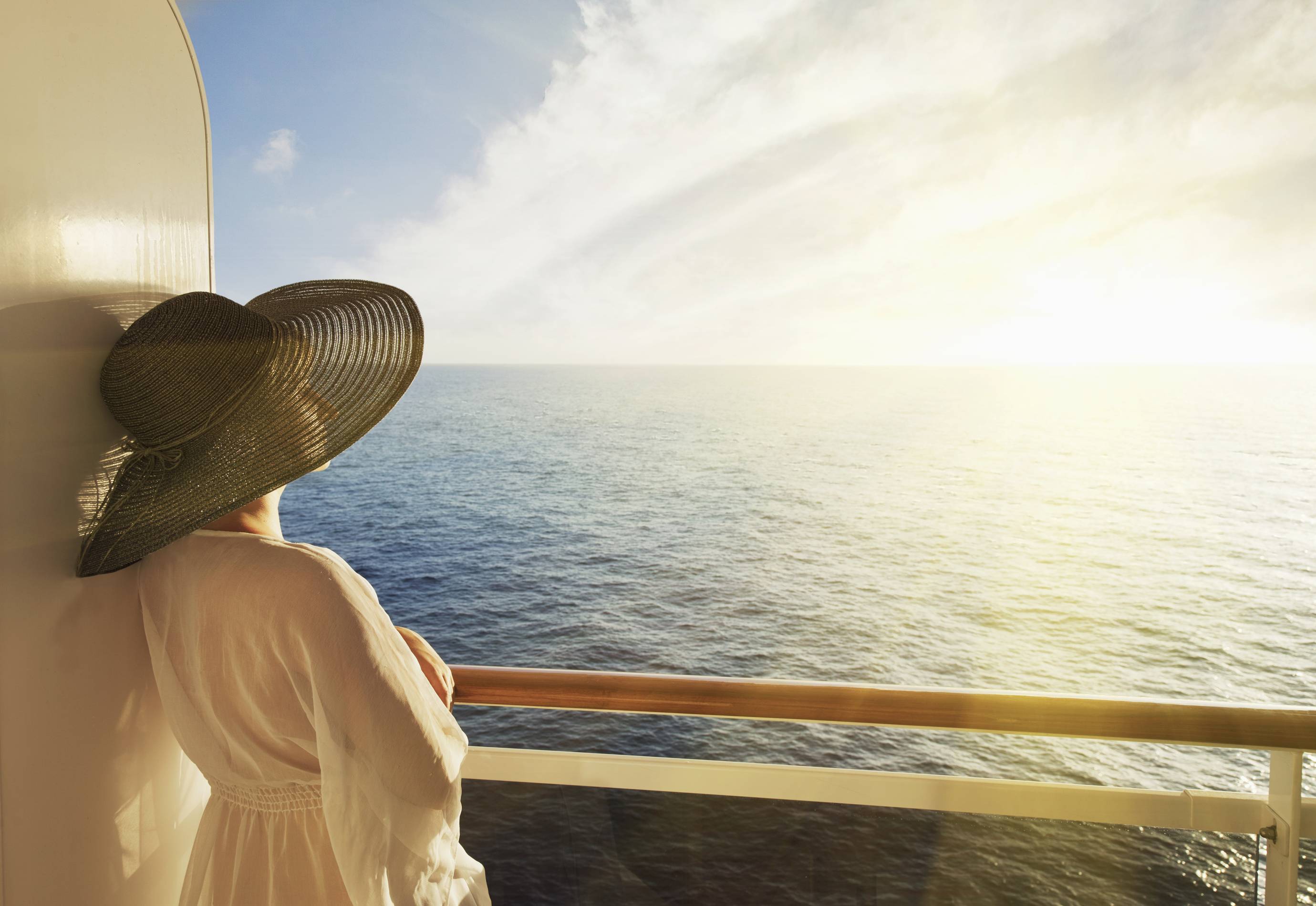 a woman in a large hat stands at the balcony of her cruise cabin