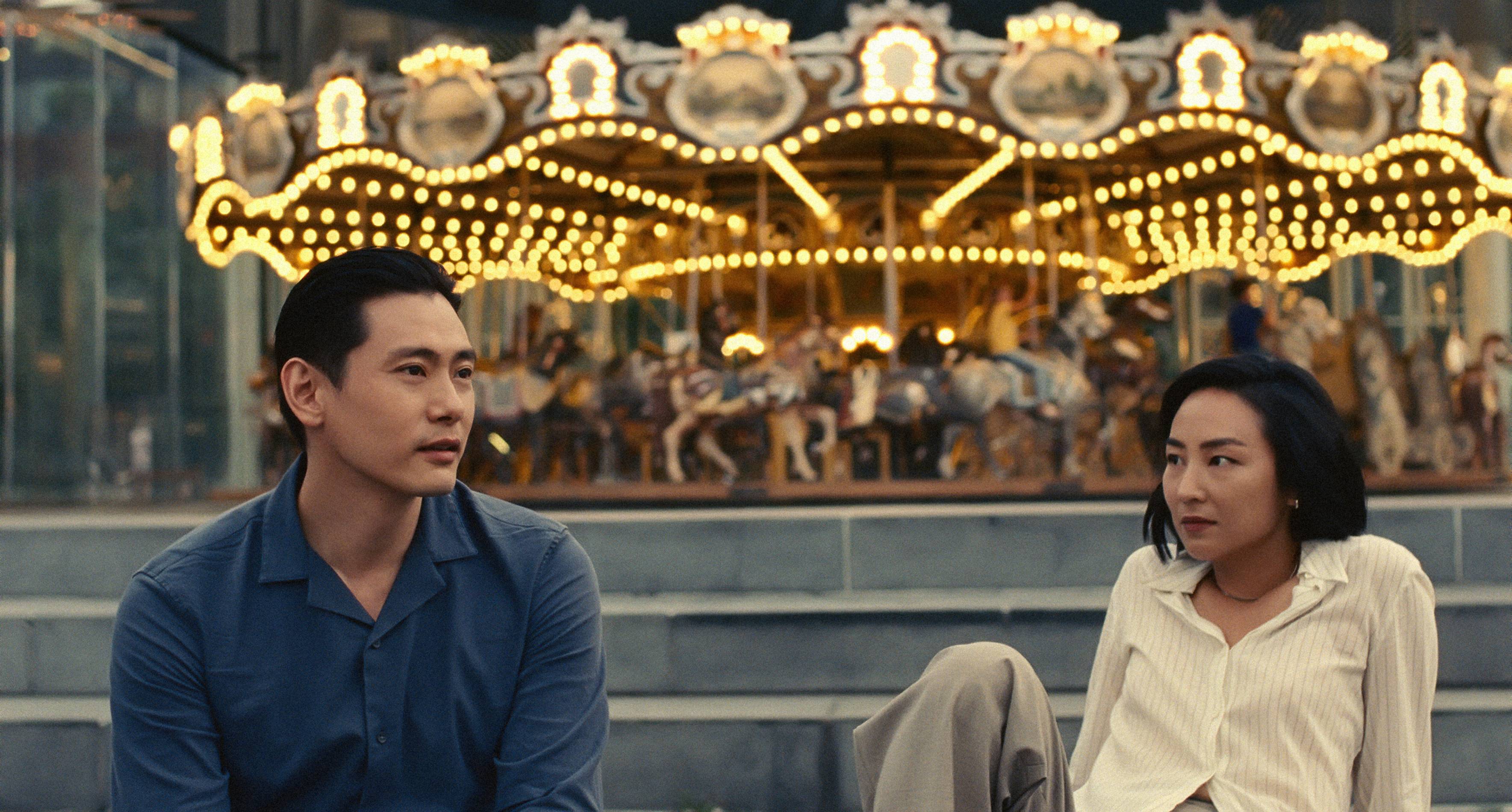 Teo Yoo and Greta Lee sit in front of a carousel