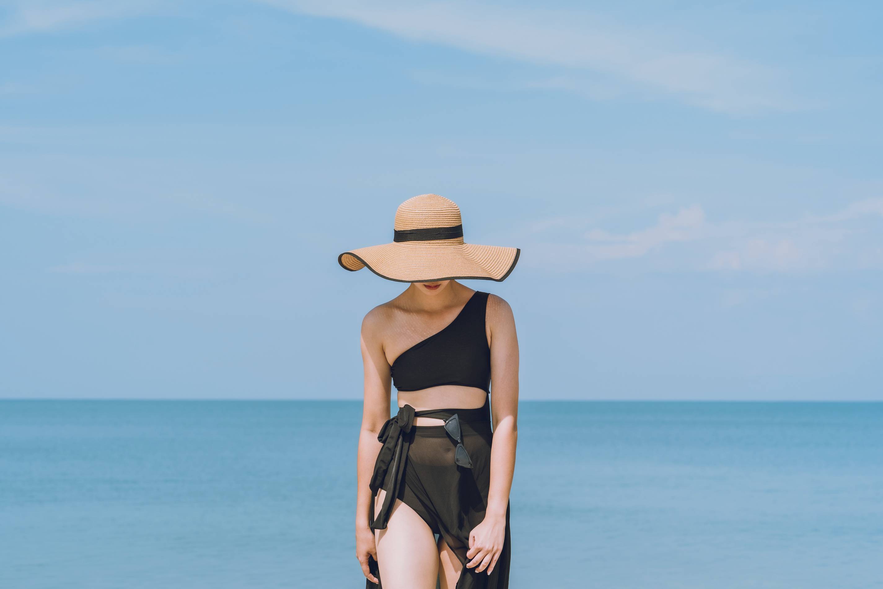 a woman walks by the sea in a black bathing suit and dress with a big hat